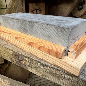 May include: A rectangular gray sharpening stone with a visible weight of 1715g written on the side, secured within a light brown wooden base featuring two parallel wooden guide rails attached to the flat platform for stability and support.