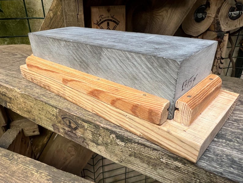 May include: A rectangular gray sharpening stone with a visible weight of 1715g written on the side, secured within a light brown wooden base featuring two parallel wooden guide rails attached to the flat platform for stability and support.