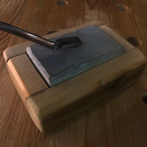 Puede incluir: Un bloque de afilado de madera con una piedra de afilar gris en el interior. Una herramienta de metal descansa sobre la piedra.