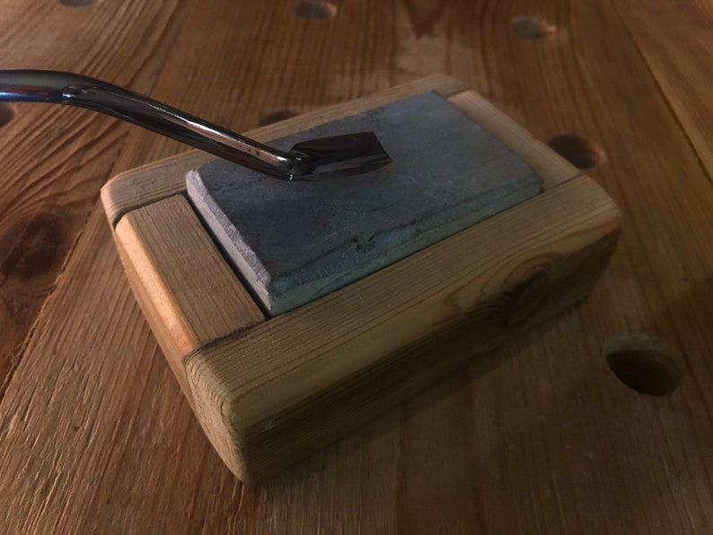 Puede incluir: Un bloque de afilado de madera con una piedra de afilar gris en el interior. Una herramienta de metal descansa sobre la piedra.