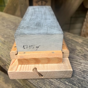 May include: A rectangular gray sharpening stone with a textured surface, featuring the handwritten text 1715 gr on its front face. The stone is securely held within a light-colored wooden base that has a visible grain and a small notch on the front.