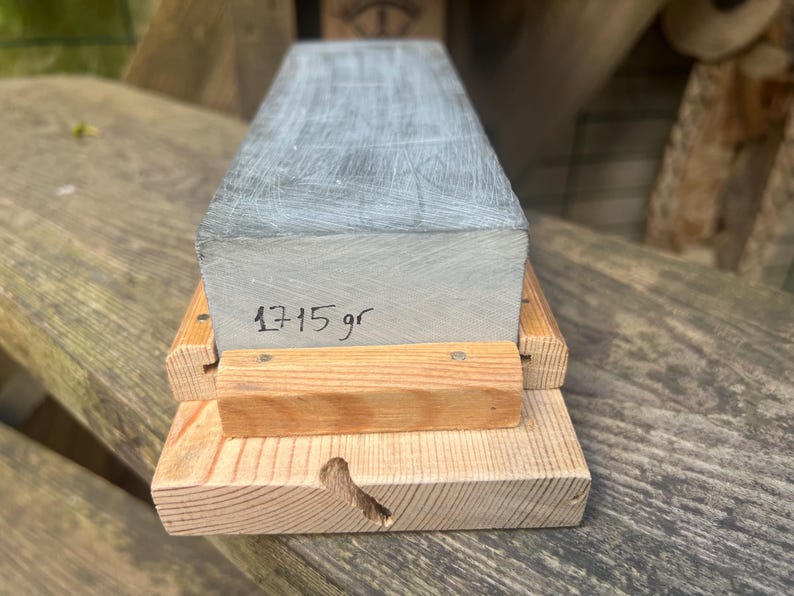 May include: A rectangular gray sharpening stone with a textured surface, featuring the handwritten text 1715 gr on its front face. The stone is securely held within a light-colored wooden base that has a visible grain and a small notch on the front.