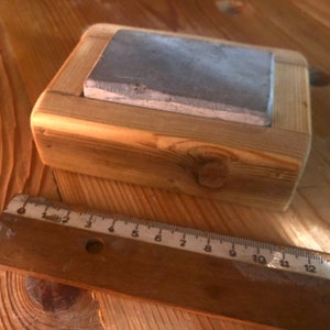 Puede incluir: Una piedra de afilar gris est&aacute; colocada en un marco de madera. El marco est&aacute; hecho de madera de corte &aacute;spero con nudos y grano visibles. La piedra es rectangular y tiene una superficie ligeramente texturizada.