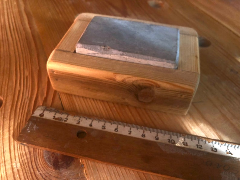 Puede incluir: Una piedra de afilar gris est&aacute; colocada en un marco de madera. El marco est&aacute; hecho de madera de corte &aacute;spero con nudos y grano visibles. La piedra es rectangular y tiene una superficie ligeramente texturizada.
