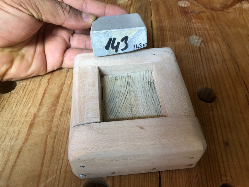 Pu&ograve; includere: Una scatola di legno con un'apertura rettangolare, progettata per contenere una pietra per affilare grigia. La pietra &egrave; etichettata "143".