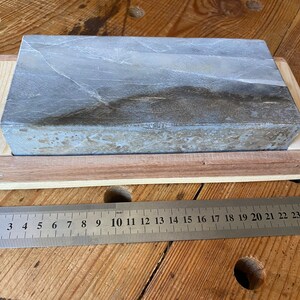 Puede incluir: Una piedra de afilar gris con una base de madera. La piedra mide aproximadamente 25 cm de largo.