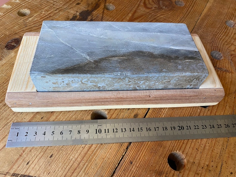 Puede incluir: Una piedra de afilar gris con una base de madera. La piedra mide aproximadamente 25 cm de largo.