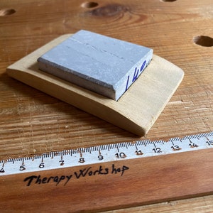 May include: A gray sharpening stone with a wooden base. The stone is rectangular and has a light gray color. The base is made of light wood and has a curved shape. The stone is labeled "142".