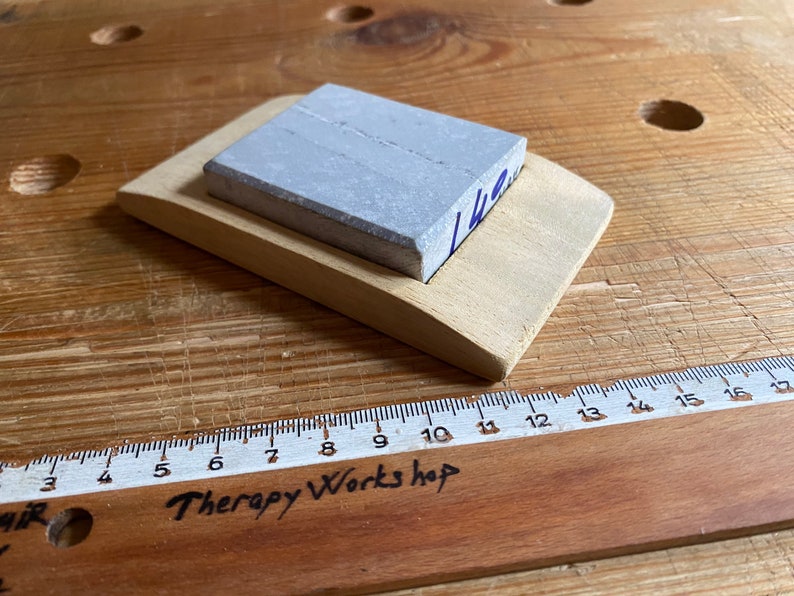May include: A gray sharpening stone with a wooden base. The stone is rectangular and has a light gray color. The base is made of light wood and has a curved shape. The stone is labeled "142".