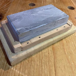 Peut inclure: Une pierre &agrave; aiguiser grise avec une base en bois. La pierre est rectangulaire et a une surface rugueuse. La base est en bois clair et comporte une s&eacute;rie de petits trous.