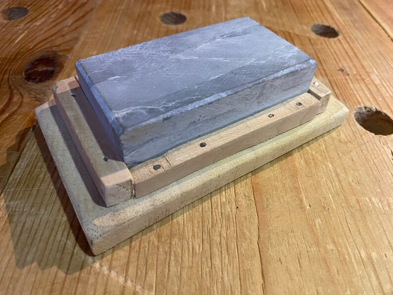 Peut inclure: Une pierre &agrave; aiguiser grise avec une base en bois. La pierre est rectangulaire et a une surface rugueuse. La base est en bois clair et comporte une s&eacute;rie de petits trous.