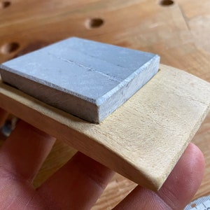 May include: A gray rectangular sharpening stone sits on a light wood base. The base is shaped like a boat and has a rounded edge.