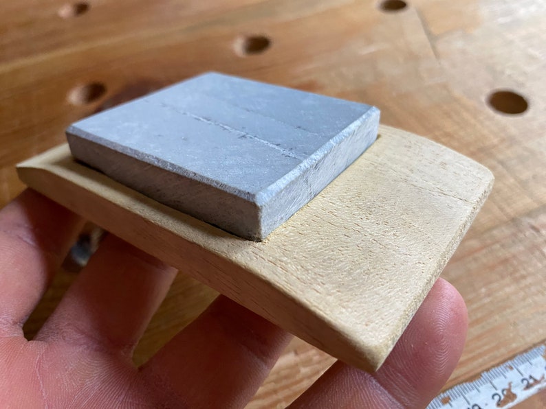 May include: A gray rectangular sharpening stone sits on a light wood base. The base is shaped like a boat and has a rounded edge.
