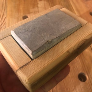 Puede incluir: Una piedra de afilar gris se encuentra en un marco de madera. El marco est&aacute; hecho de madera clara y tiene un dise&ntilde;o simple.