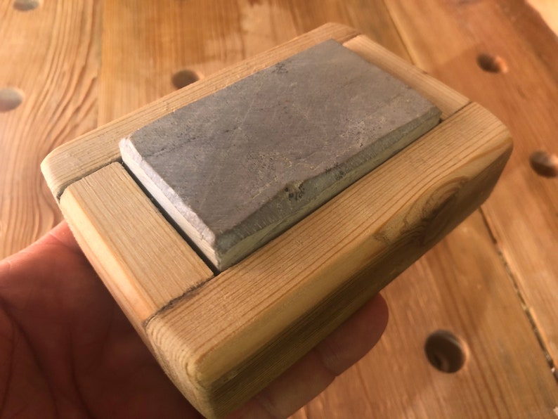 Puede incluir: Una piedra de afilar gris se encuentra en un marco de madera. El marco est&aacute; hecho de madera clara y tiene un dise&ntilde;o simple.