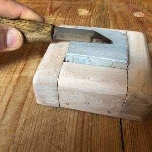 May include: A wooden sharpening block with a gray sharpening stone. A hand is holding a knife and sharpening it on the stone.