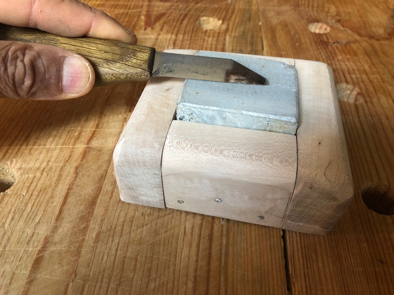 Pu&ograve; includere: Un blocco di affilatura in legno con una pietra di affilatura grigia. Una mano tiene un coltello e lo affila sulla pietra.