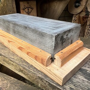 May include: A rectangular gray stone sharpening block mounted on a light-colored wooden base. The base features two parallel wooden guide rails secured with small metal fasteners, designed to hold the stone in a fixed position during use.