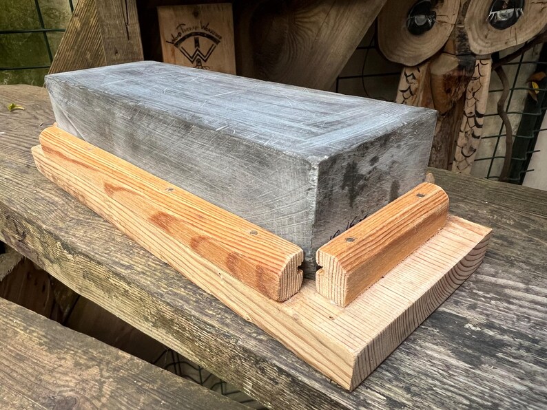 May include: A rectangular gray stone sharpening block mounted on a light-colored wooden base. The base features two parallel wooden guide rails secured with small metal fasteners, designed to hold the stone in a fixed position during use.