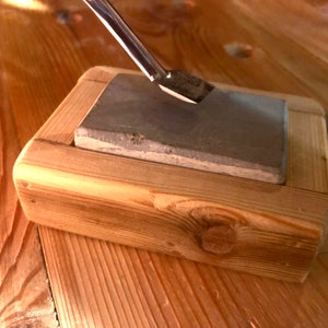 Puede incluir: Primer plano de una piedra de afilar en un soporte de madera. Una herramienta de metal se est&aacute; afilando en la piedra.