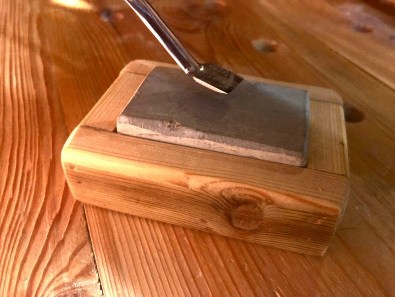 Puede incluir: Primer plano de una piedra de afilar en un soporte de madera. Una herramienta de metal se est&aacute; afilando en la piedra.