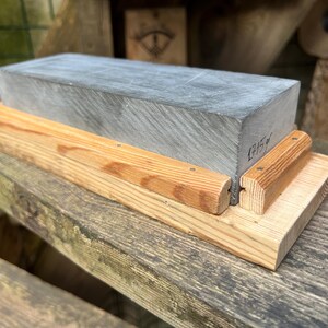 May include: A rectangular gray sharpening stone mounted on a light-colored wooden base. The stone features a visible handwritten inscription on its side that reads 1715g. The wooden base consists of a flat bottom piece with two side rails.