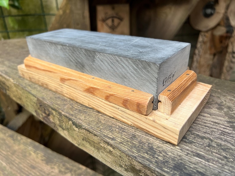 May include: A rectangular gray sharpening stone mounted on a light-colored wooden base. The stone features a visible handwritten inscription on its side that reads 1715g. The wooden base consists of a flat bottom piece with two side rails.