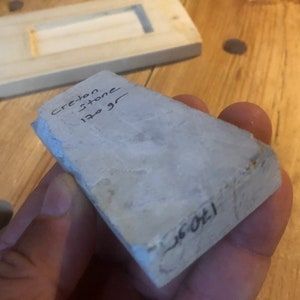 May include: A light grey stone with the words "Creston Stone 130 gr" written on it. The stone is being held in a hand.