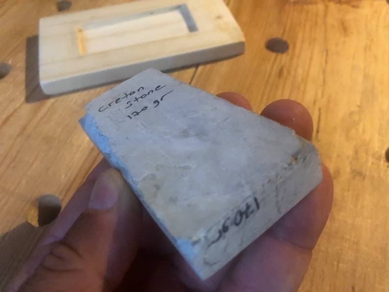 May include: A light grey stone with the words "Creston Stone 130 gr" written on it. The stone is being held in a hand.
