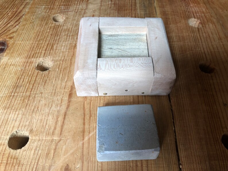 Pu&ograve; includere: Una scatola di legno con un sasso grigio all'interno. La scatola &egrave; fatta di legno chiaro e ha un design semplice. Il sasso &egrave; rettangolare e ha una texture ruvida.