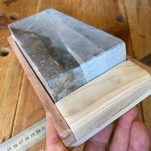 Puede incluir: Una piedra de afilar gris y blanca en un soporte de madera. La piedra es rectangular y tiene una superficie rugosa. El soporte es de madera clara y tiene un dise&ntilde;o sencillo.