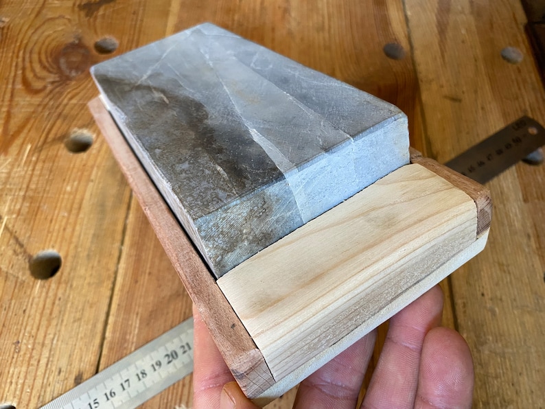 Puede incluir: Una piedra de afilar gris y blanca en un soporte de madera. La piedra es rectangular y tiene una superficie rugosa. El soporte es de madera clara y tiene un dise&ntilde;o sencillo.