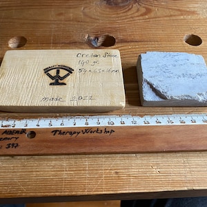 May include: A wooden block with the text "Cretan Stone 140 gr 54&times;63&times;16m made 2022" and a logo for "Therapy Workshop". A gray stone measuring 5.4 cm x 6.3 cm x 1.6 cm is next to the block. A wooden ruler with the text "SAVAŞ ALDEMİR 1978 Memory 5A; No: 397 Therapy Workshop" is below the block and stone.