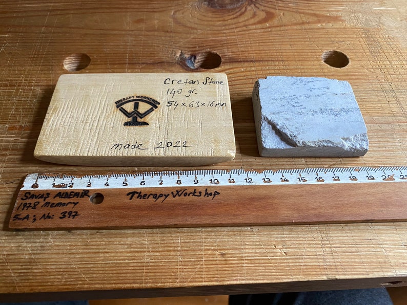 May include: A wooden block with the text "Cretan Stone 140 gr 54&times;63&times;16m made 2022" and a logo for "Therapy Workshop". A gray stone measuring 5.4 cm x 6.3 cm x 1.6 cm is next to the block. A wooden ruler with the text "SAVAŞ ALDEMİR 1978 Memory 5A; No: 397 Therapy Workshop" is below the block and stone.