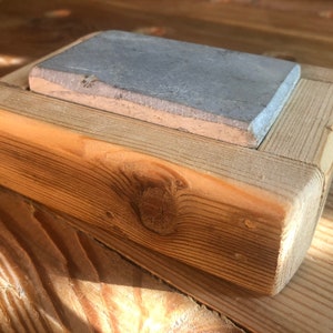 Puede incluir: Una piedra de afilar rectangular gris se encuentra en un marco de madera. El marco est&aacute; hecho de madera toscamente tallada con un acabado natural.