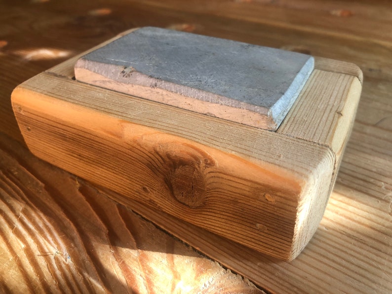 Puede incluir: Una piedra de afilar rectangular gris se encuentra en un marco de madera. El marco est&aacute; hecho de madera toscamente tallada con un acabado natural.