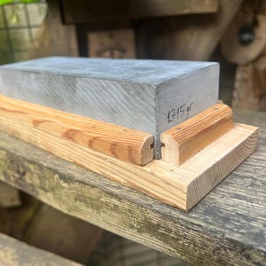 May include: A rectangular gray sharpening stone set within a light-colored wooden base. The stone features a smooth, flat top surface and a side face with the handwritten text 1715 gr. The wooden base consists of a flat bottom piece and two side rails.