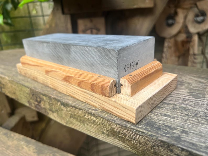 May include: A rectangular gray sharpening stone set within a light-colored wooden base. The stone features a smooth, flat top surface and a side face with the handwritten text 1715 gr. The wooden base consists of a flat bottom piece and two side rails.
