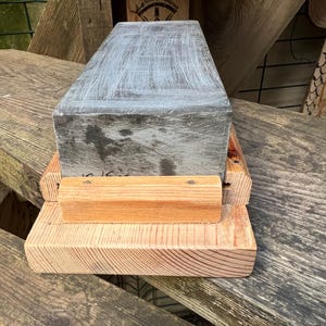 May include: A rectangular gray sharpening stone with visible surface scratches, secured within a light-colored wooden base. The stone is positioned horizontally, featuring a flat top surface and a natural wood grain texture on the supporting frame.