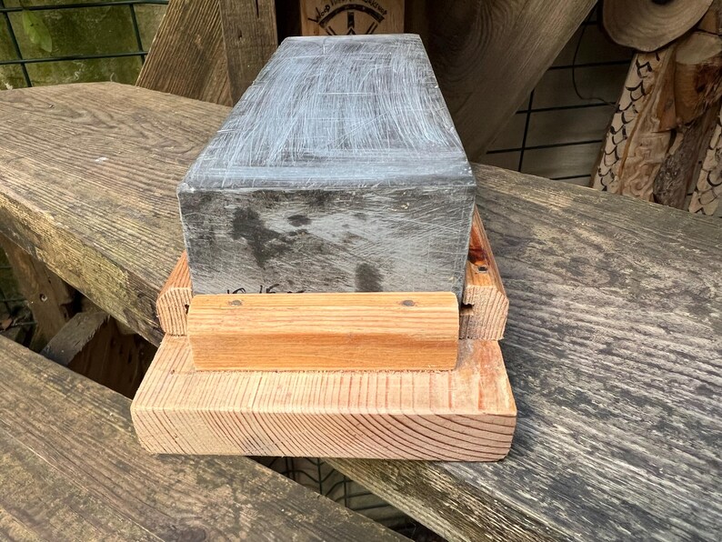May include: A rectangular gray sharpening stone with visible surface scratches, secured within a light-colored wooden base. The stone is positioned horizontally, featuring a flat top surface and a natural wood grain texture on the supporting frame.