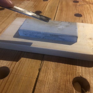 May include: A grey sharpening stone sits on a wooden base. A metal tool is being sharpened on the stone.