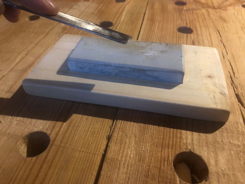 May include: A grey sharpening stone sits on a wooden base. A metal tool is being sharpened on the stone.