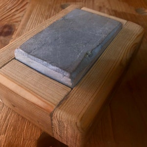 Puede incluir: Una piedra de afilar gris est&aacute; colocada en un marco de madera. El marco est&aacute; hecho de madera marr&oacute;n claro y tiene un dise&ntilde;o simple.