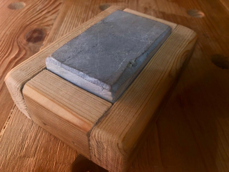 Puede incluir: Una piedra de afilar gris est&aacute; colocada en un marco de madera. El marco est&aacute; hecho de madera marr&oacute;n claro y tiene un dise&ntilde;o simple.