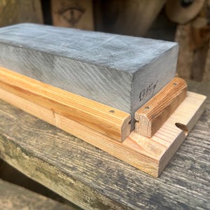 May include: A rectangular gray sharpening stone with the text 1715 gr written on the side. The stone is held in place by a light-colored wooden base featuring two parallel wooden rails secured with small metal nails along the sides of the stone.
