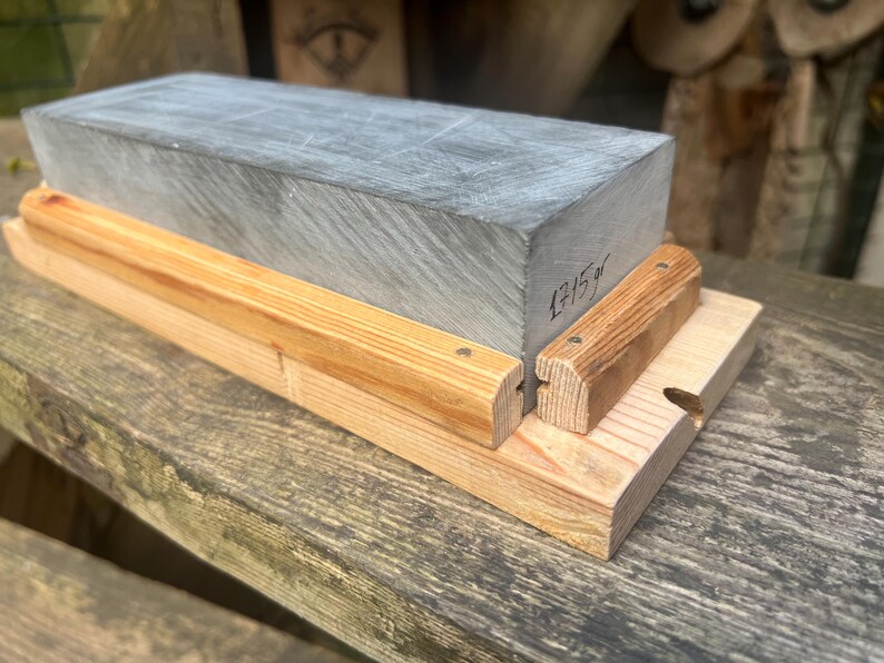 May include: A rectangular gray sharpening stone with the text 1715 gr written on the side. The stone is held in place by a light-colored wooden base featuring two parallel wooden rails secured with small metal nails along the sides of the stone.