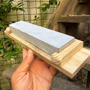 May include: A rectangular light gray sharpening stone mounted on a light-colored wooden base. The base features raised wooden edges on the sides to secure the stone in place. The sharpening stone has a flat, textured surface and a uniform color.