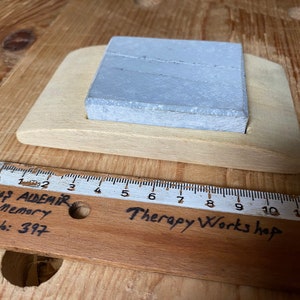 May include: A gray rectangular sharpening stone sits in a wooden holder. The holder is labeled "Therapy Works hop". A wooden ruler with the text "SAVAŞ ALDEMİR 1978 Memory 5-A No: 397" is in the foreground.