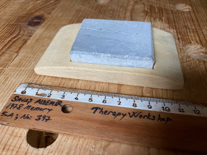 May include: A gray rectangular sharpening stone sits in a wooden holder. The holder is labeled "Therapy Works hop". A wooden ruler with the text "SAVAŞ ALDEMİR 1978 Memory 5-A No: 397" is in the foreground.