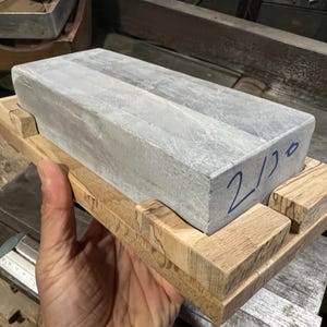 May include: A rectangular gray sharpening stone with the number "2/10" written in blue ink, resting on a wooden base. The stone is held in a hand, showcasing its size and texture. The wooden base is made of light brown wood.